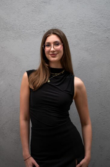 Young woman with long brown hair wearing round glasses and a black sleeveless dress, standing against a gray textured wall.