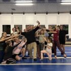 IMG_7561 Duży - Fit&More Group of kids and a coach posing playfully on blue gym mats in a training hall, forming a cheer pose.