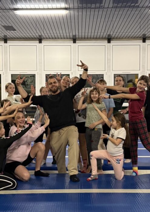 IMG_7561 Duży - Fit&More Group of kids and a coach posing playfully on blue gym mats in a training hall, forming a cheer pose.