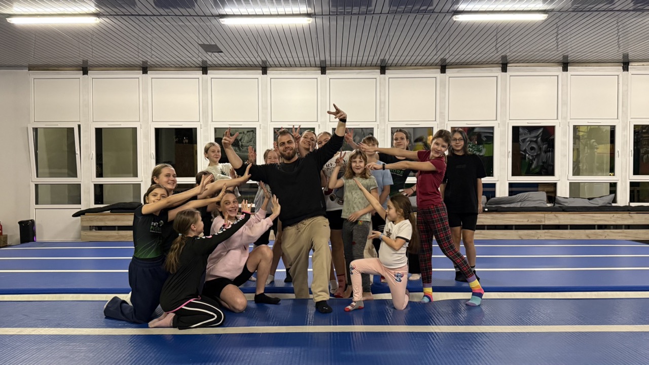 Group of kids and a coach posing playfully on blue gym mats in a training hall, forming a cheer pose.