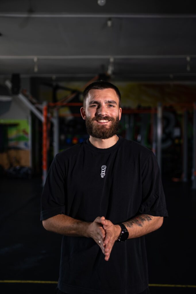 Smiling man with a beard in a black t-shirt standing in a gym, hands clasped in front of him.