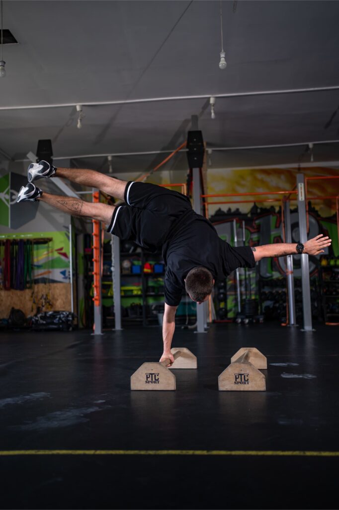 Athlete mid-handstand in a gym, balancing on one hand with legs extended sideways above wooden blocks labeled PT SPORT in the foreground.
