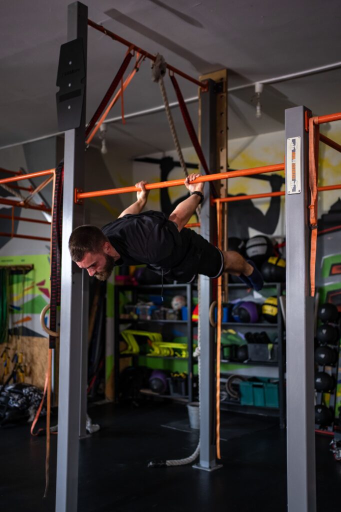 Man in a dark shirt hanging horizontally from an orange gym bar on a metal frame during a workout in a gym.