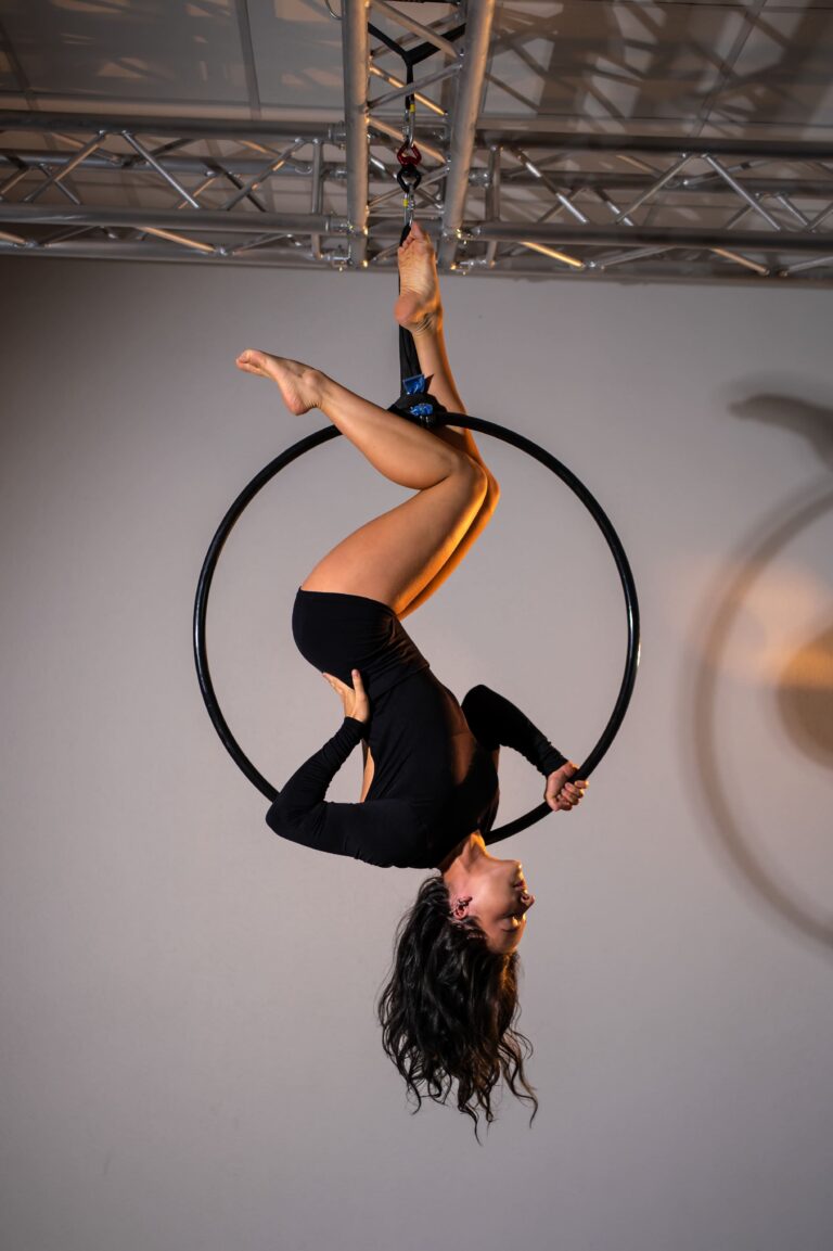 Two aerialists in black perform on a suspended hoop; one hangs inverted inside the hoop while the other supports from below in a studio setting.