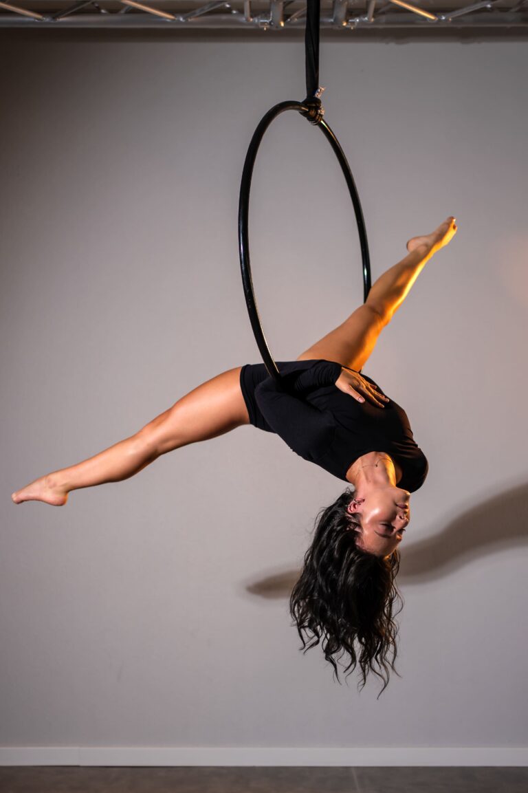A performer in black shorts on an aerial hoop, upside down with one leg extended outward.