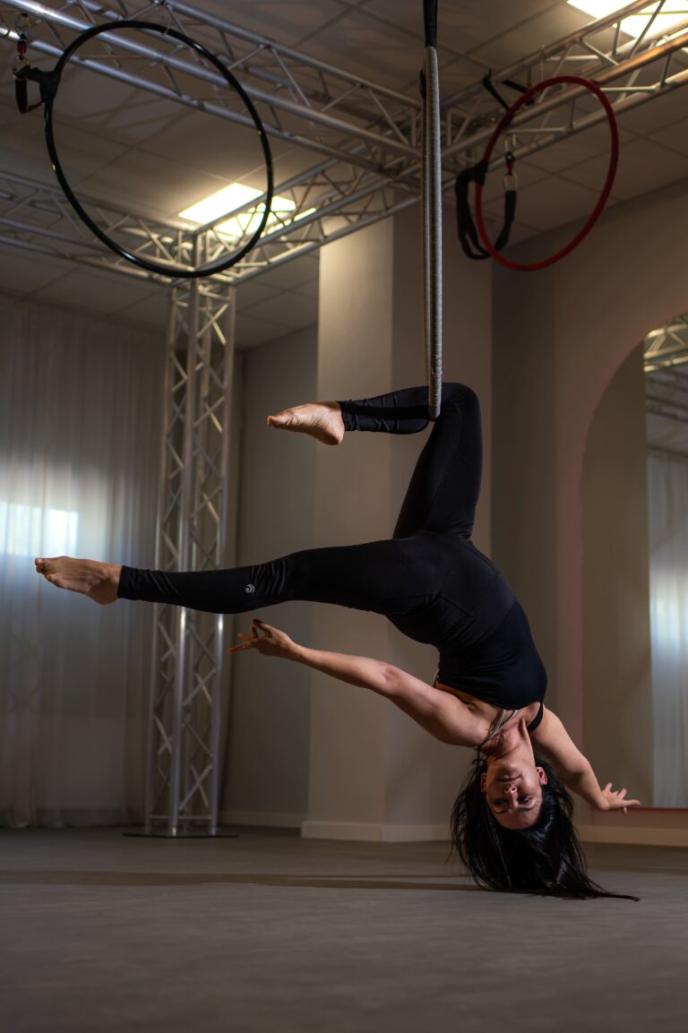 Aerialist hanging upside down from a rope, legs wrapped around it in a black athletic outfit inside a studio with rigging overhead.