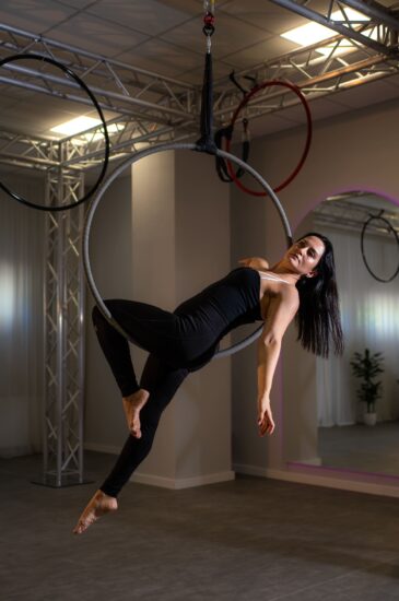 Woman in black athletic wear performs on an aerial hoop suspended from a rig in a studio, hanging mid-air with legs extended behind.