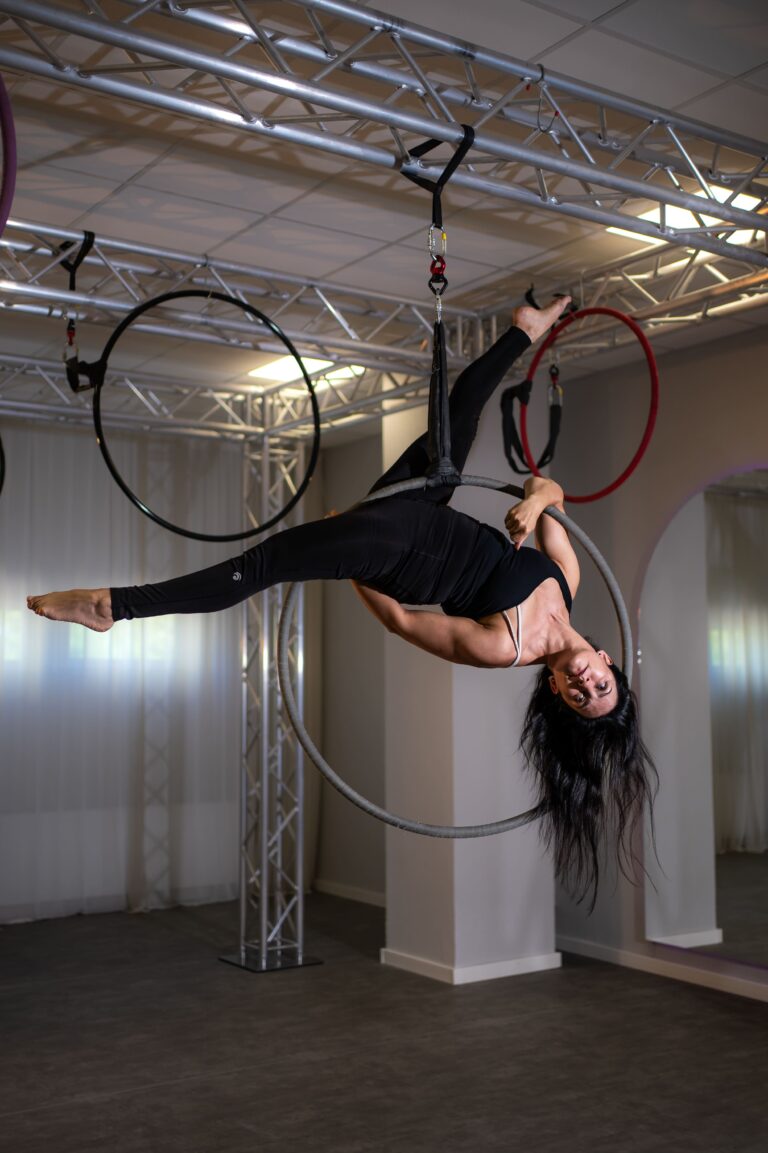 Aerialist in black performs on a suspended hoop, hanging upside down from a silver truss in a studio.