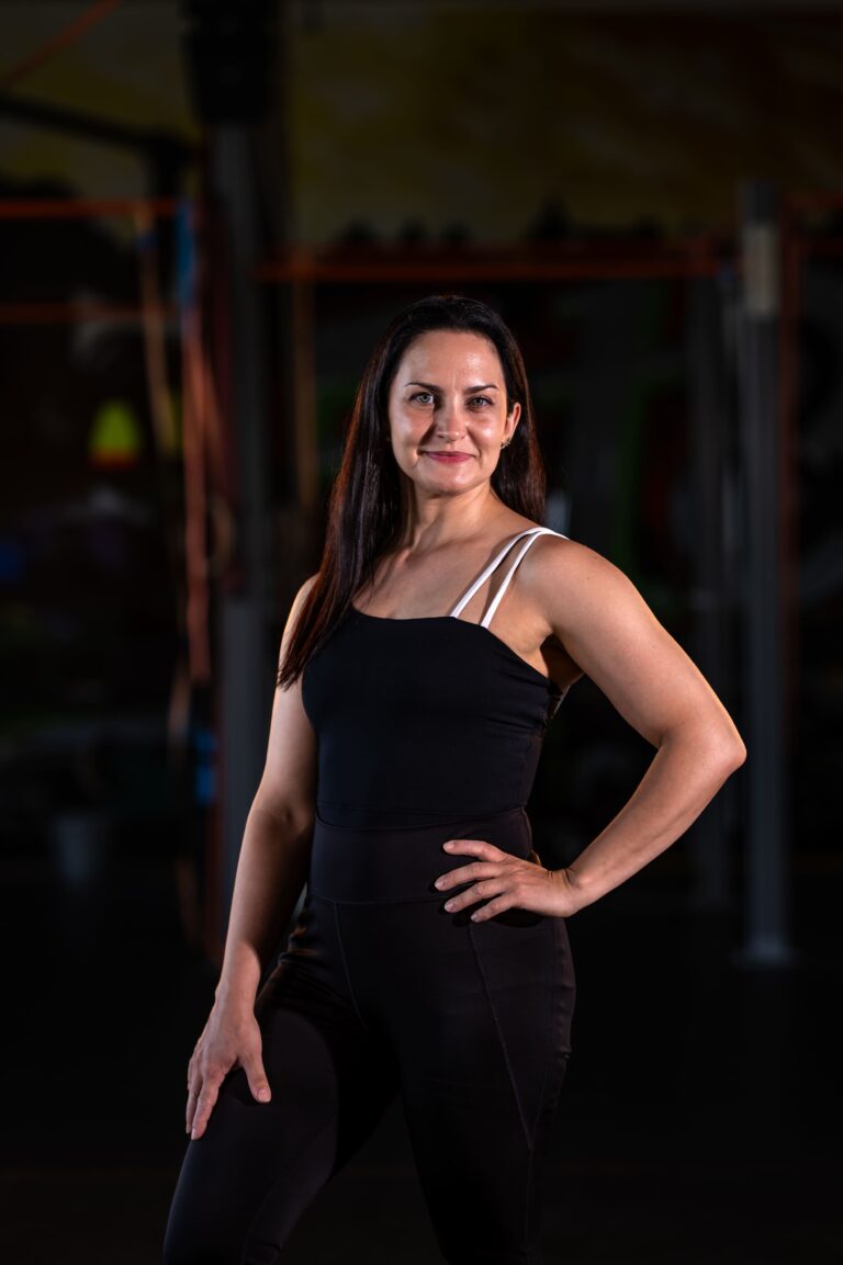 Woman in a black athletic outfit standing with hand on hip in a dimly lit gym backdrop with equipment blurred behind her