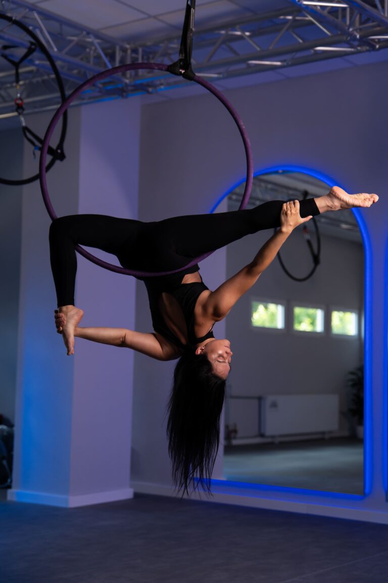 Aerialist in black grips a purple aerial hoop, hanging upside down with legs extended in a lit indoor gym.