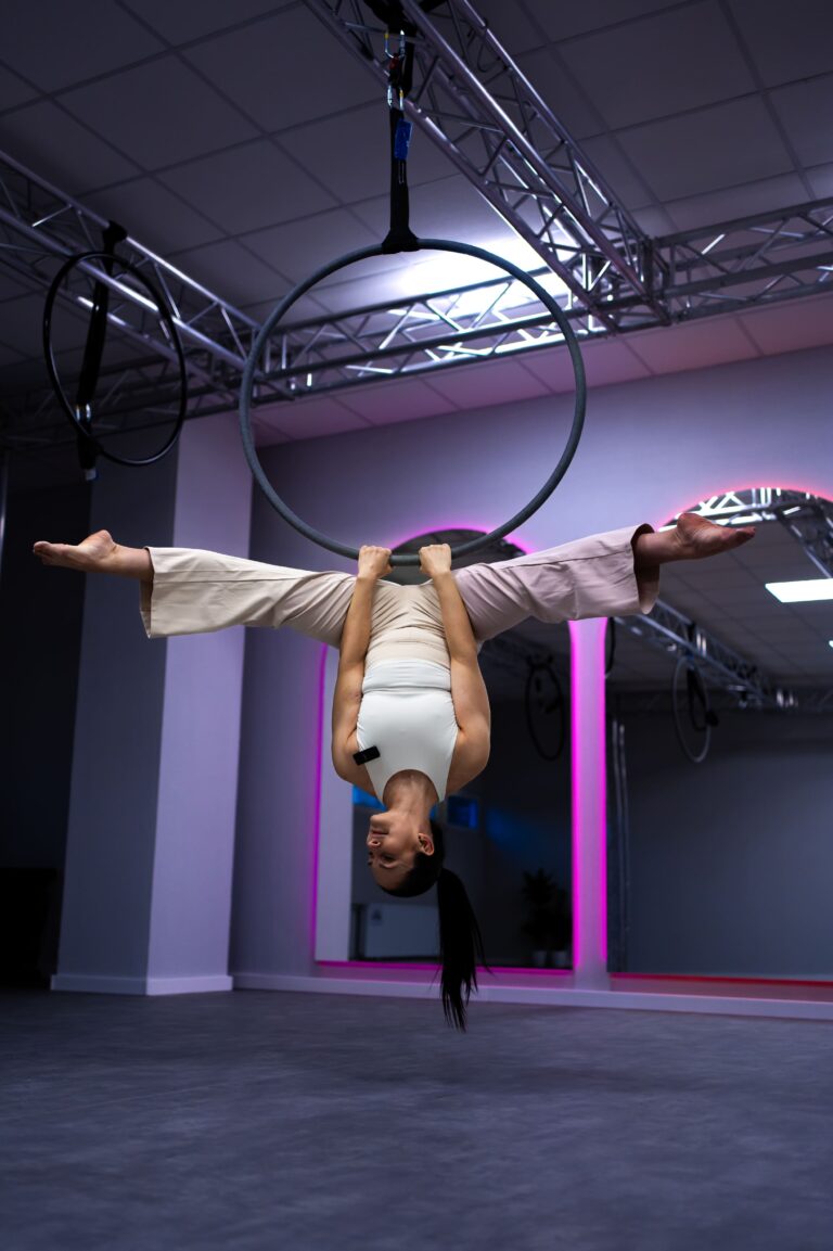 Aerialist in white costume hangs upside down, gripping a circular hoop suspended from the rig in a neon-lit studio.