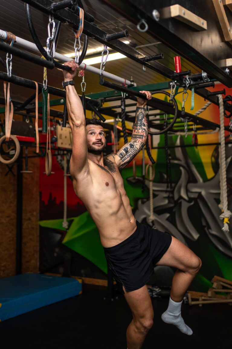 Man shirtless with tattoos hanging from gym bars in an obstacle course gym, performing a pull-up motion.