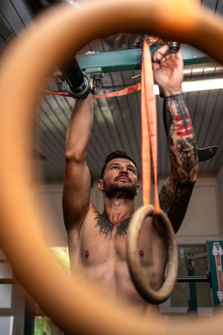Shirtless tattooed man lifts himself on gym rings overhead during a workout in a gym. A blurred orange strap dominates the foreground.