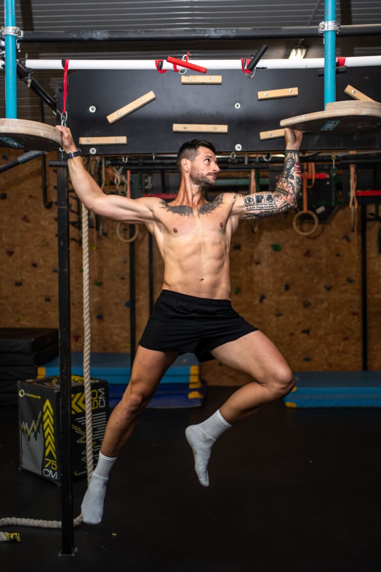 Shirtless tattooed man performing a CrossFit-style move on a metal rig, gripping two large circular wooden plates above his head.
