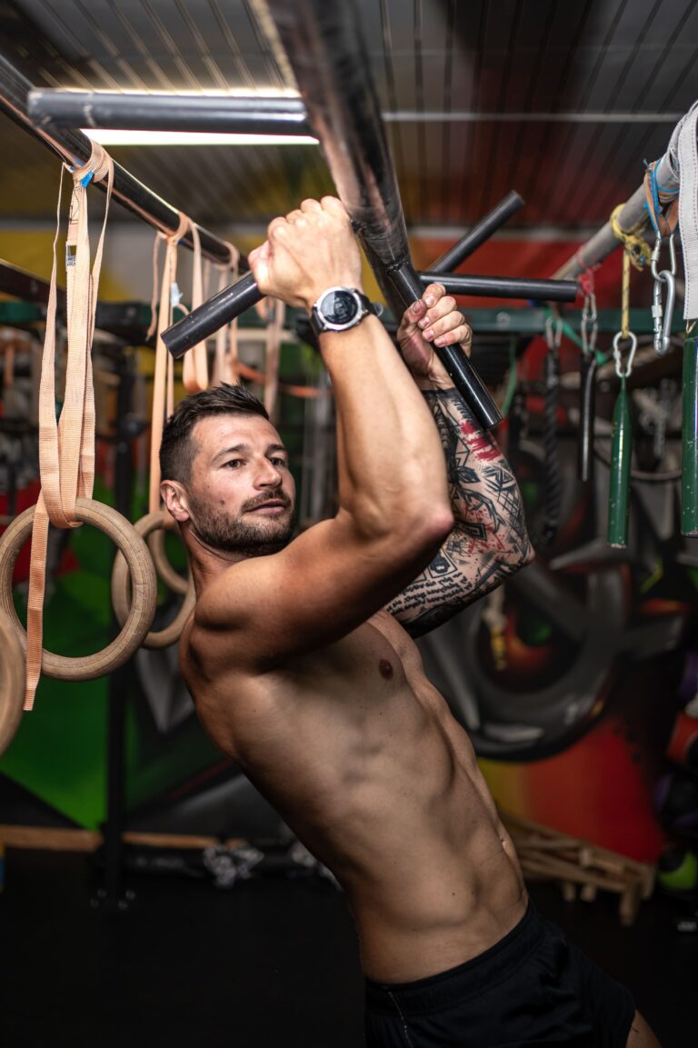Shirtless man lifting a black metal bar overhead in a colorful gym, tattoos visible on his arm, rings and straps in the background.