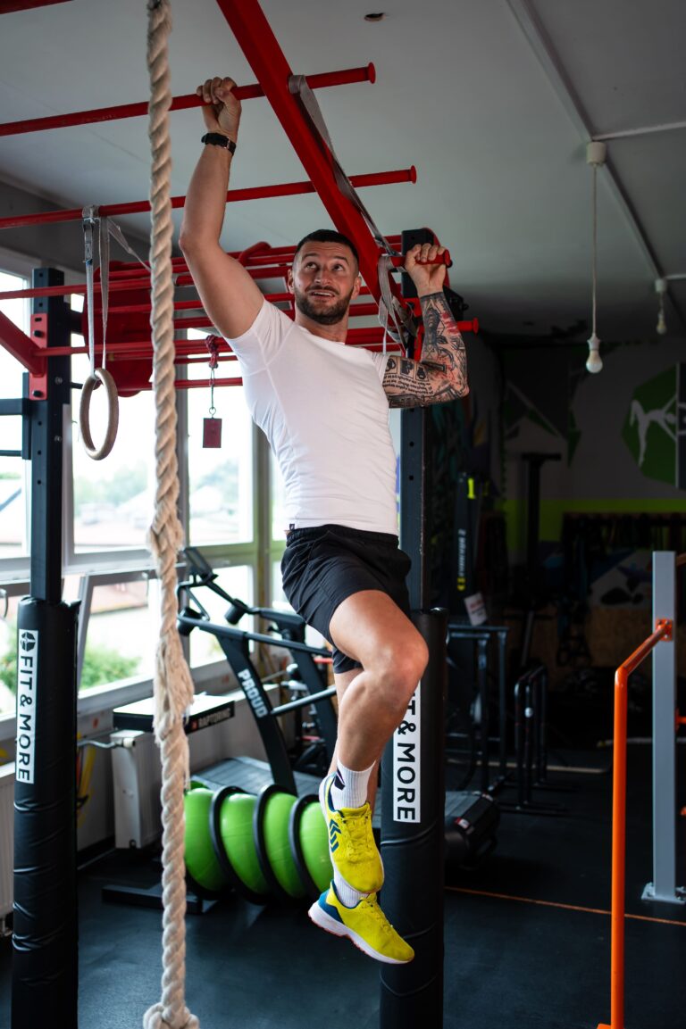 Man in white t-shirt and shorts climbing a rope in a gym, gripping red bars with tattoos on his arm and neon yellow shoes visible