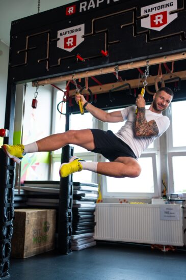 Athlete in a white shirt swinging from a black obstacle rig with yellow shoes visible, mid-air during a gym workout on hanging grips.