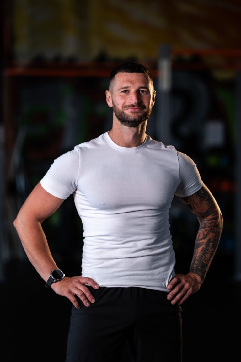 Man in a white T-shirt and black pants stands in a gym with hands on hips; visible tattoo sleeve on the right arm.