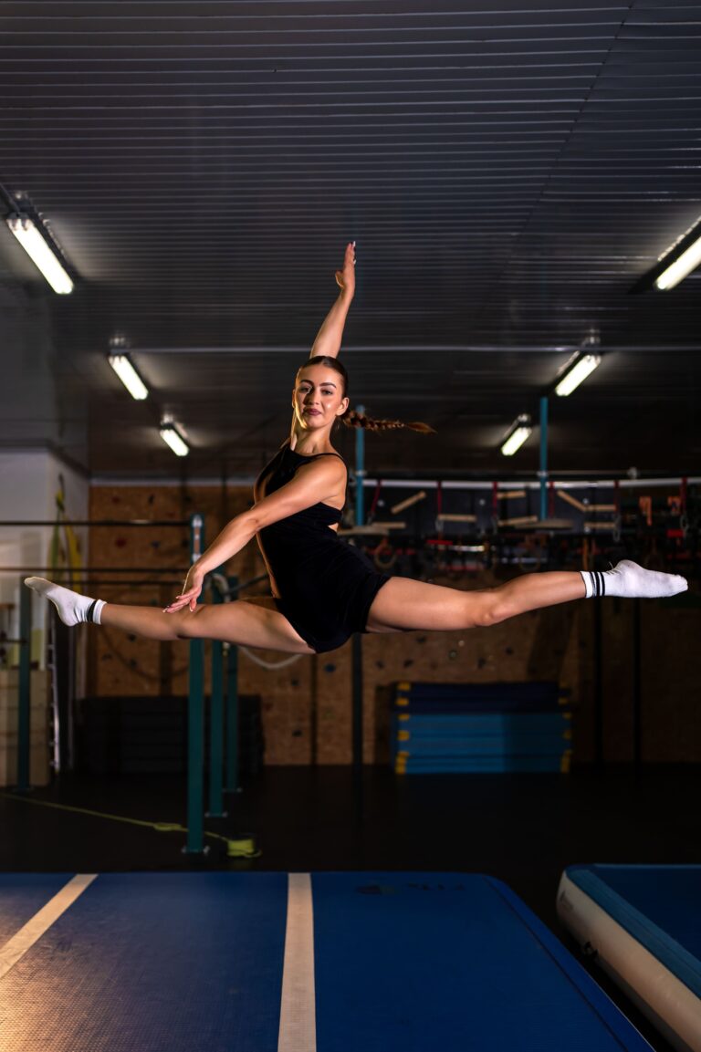 Female gymnast in a black leotard performing a full split leap in a gym, with blue mats and equipment in the background.