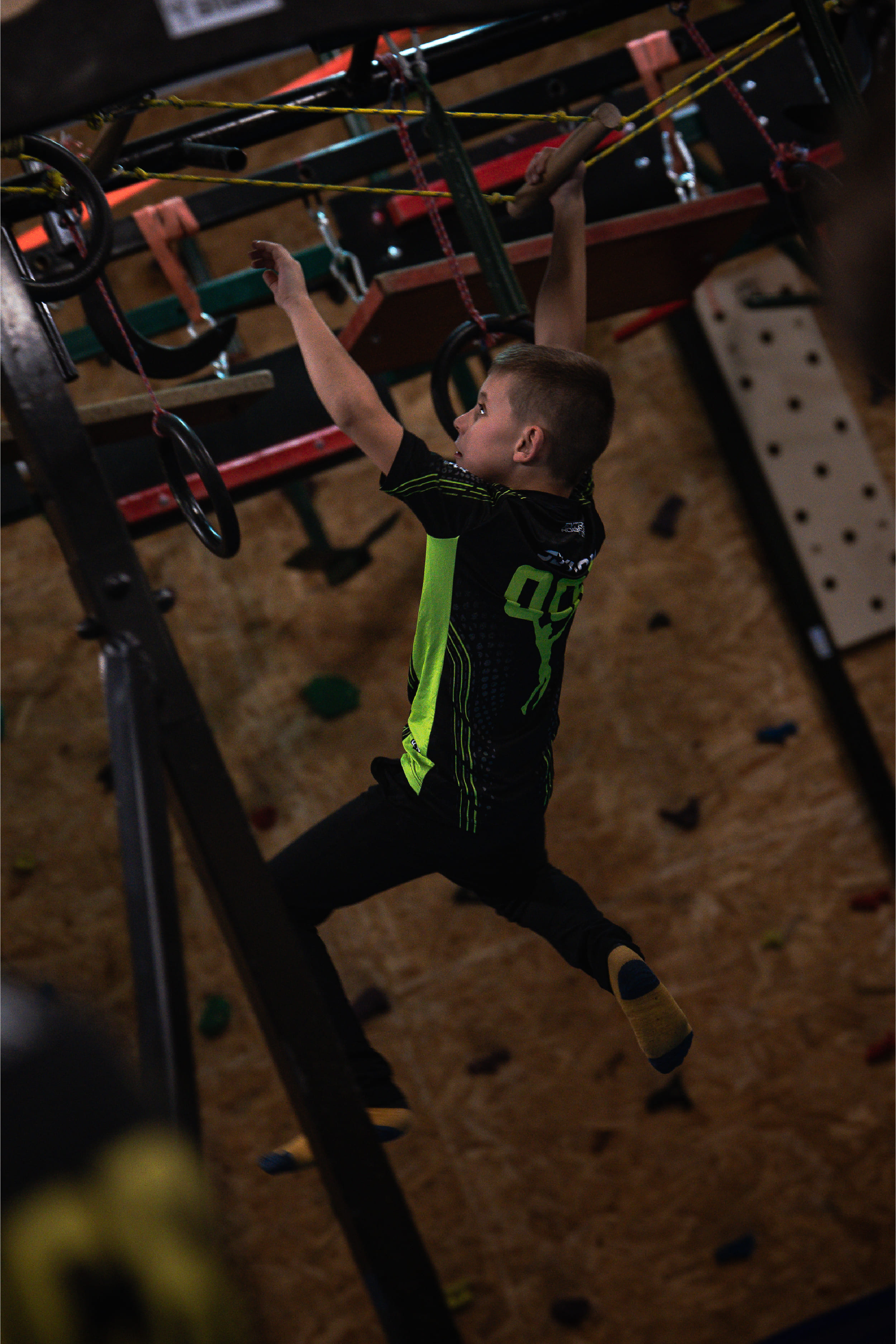 Boy in a bright green and black shirt reaches for a handhold on an indoor climbing structure with ropes and rings above him.
