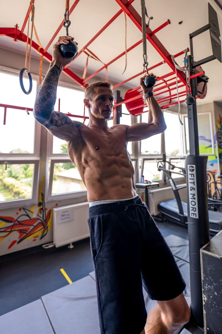 Shirtless man with tattoos lifts two black kettlebells overhead in a gym with red metal rigging and equipment nearby.