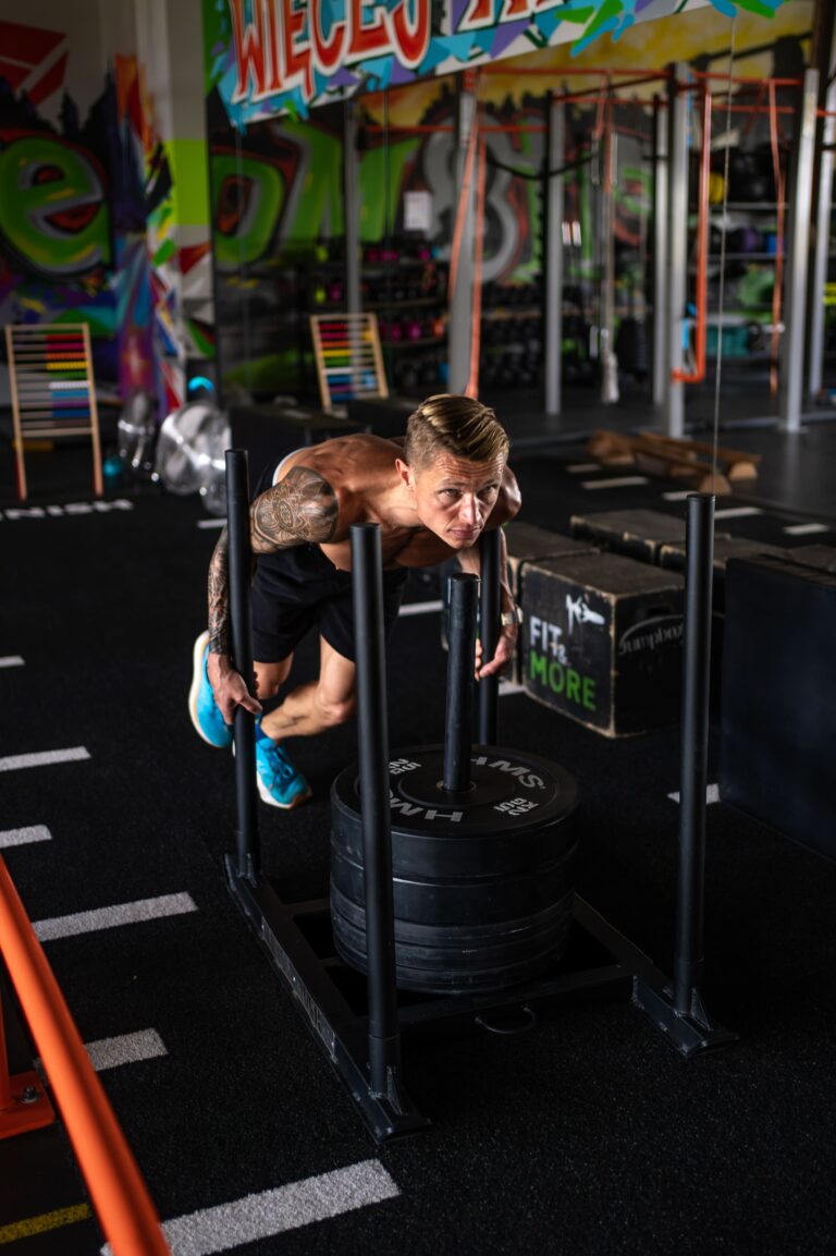 Shirtless man with tattoos doing push-ups on a weight sled in a colorful gym setting.