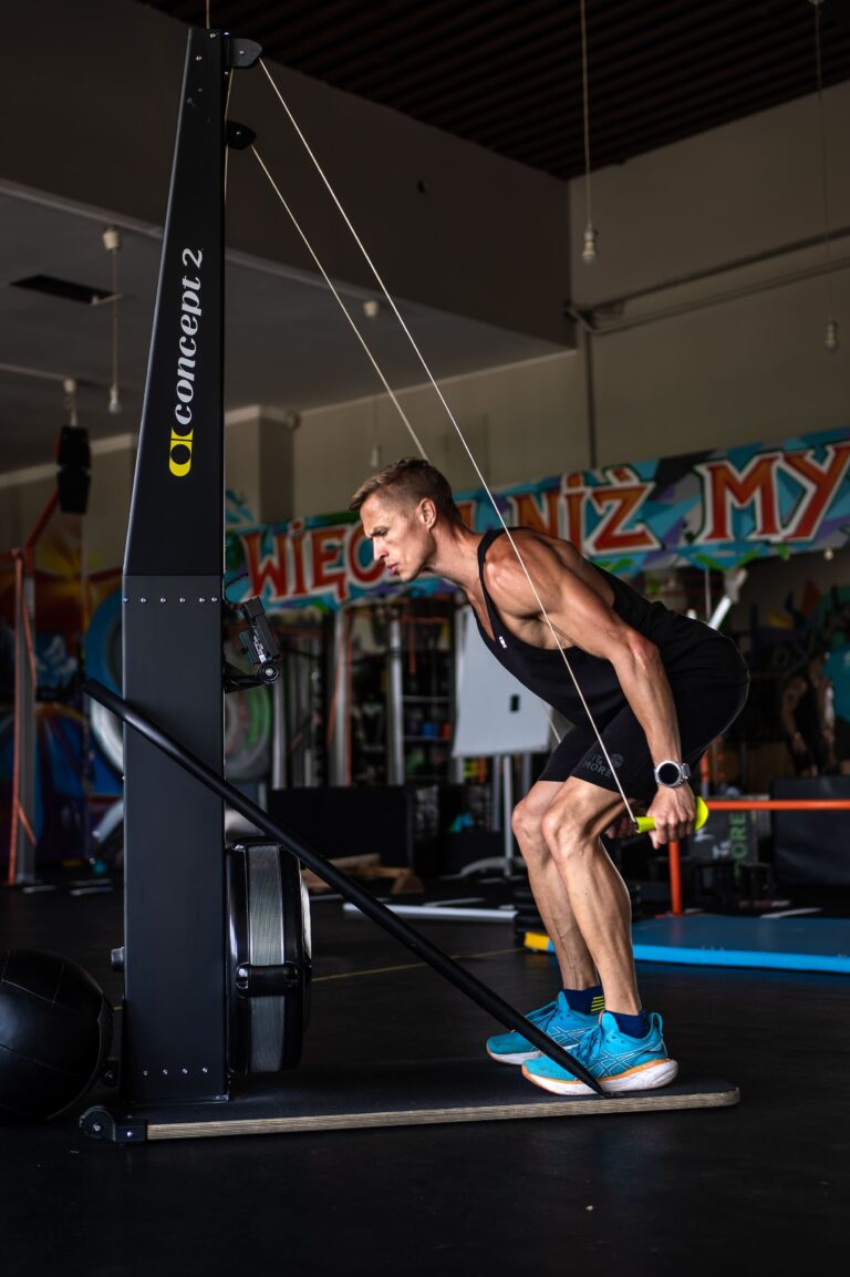 Man in a black tank top bends forward, pulling on a cable resistance machine in a gym with colorful banners in the background.