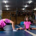Fitmore 5 wrzesnia-77 Średni - Fit&More Two young girls in pink shirts stretch on a blue gym mat; one backbends while the other sits in a full split, with gym equipment visible in the background.