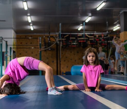 Fitmore 5 wrzesnia-77 Średni - Fit&More Two young girls in pink shirts stretch on a blue gym mat; one backbends while the other sits in a full split, with gym equipment visible in the background.