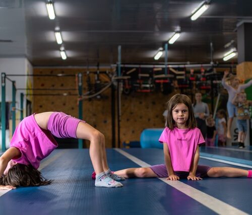 Two young girls in pink shirts stretch on a blue gym mat; one backbends while the other sits in a full split, with gym equipment visible in the background.