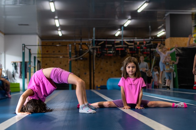 Two young girls in pink shirts stretch on a blue gym mat; one backbends while the other sits in a full split, with gym equipment visible in the background.