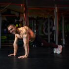 Shirtless man doing a push-up variation on a dark gym floor, tattoos visible on his arm, graffiti-covered walls in the background.