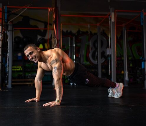 Shirtless man doing a push-up variation on a dark gym floor, tattoos visible on his arm, graffiti-covered walls in the background.