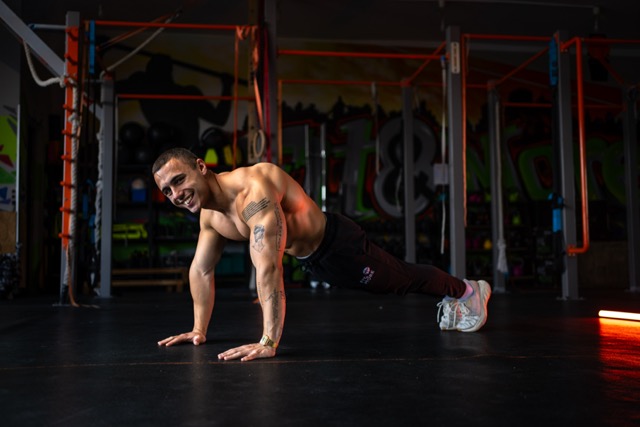 Shirtless man doing a push-up variation on a dark gym floor, tattoos visible on his arm, graffiti-covered walls in the background.