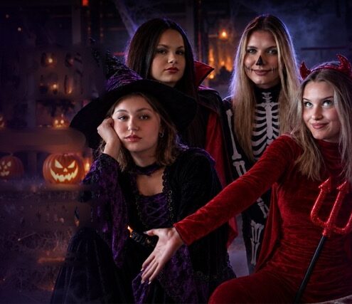 halloween_bez_napisow_3x2 Średni - Fit&More Four friends in Halloween costumes pose together at night among glowing jack-o'-lanterns; one wears a red devil outfit with a trident, others as witches and a skeleton-themed look.